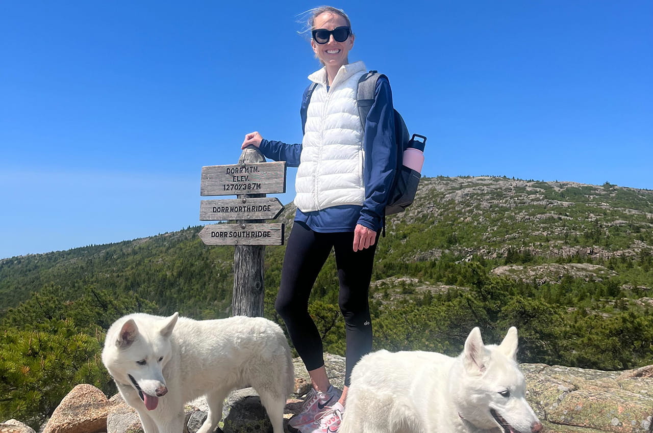 Emily McCann stands on Dorr Mountain next to a marker with her dogs.