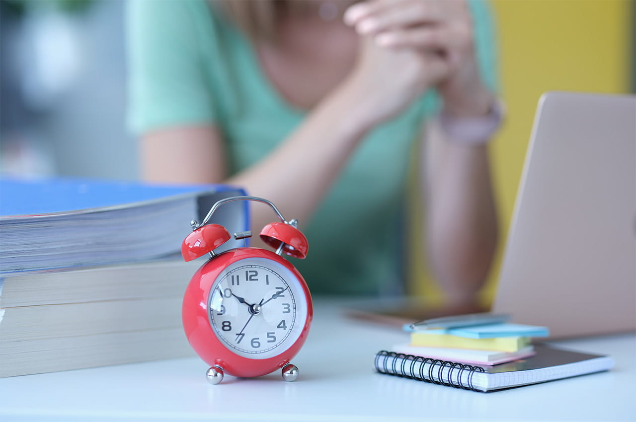 A red alarm clock is in front of an educator sitting contemplatively at her laptop.