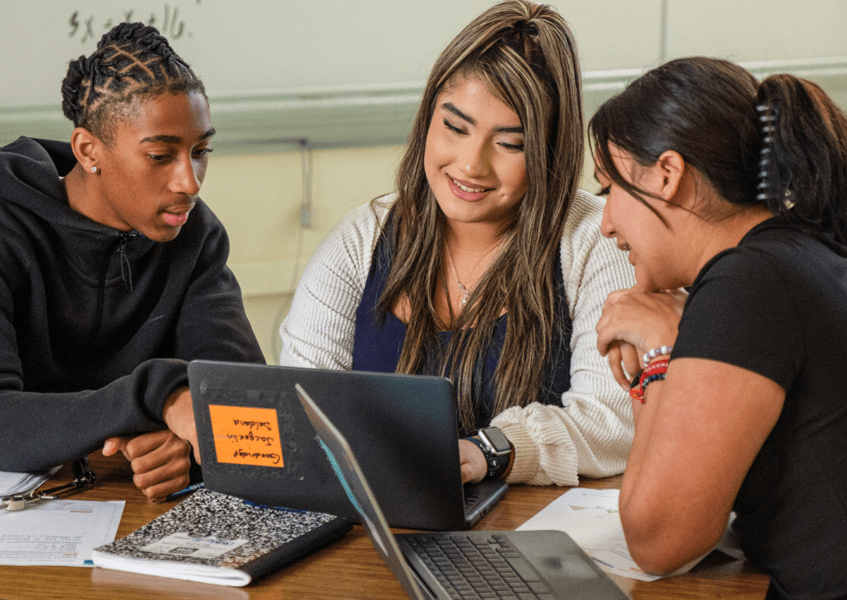 A small group of students is gathered around, looking at one of the student's laptops.