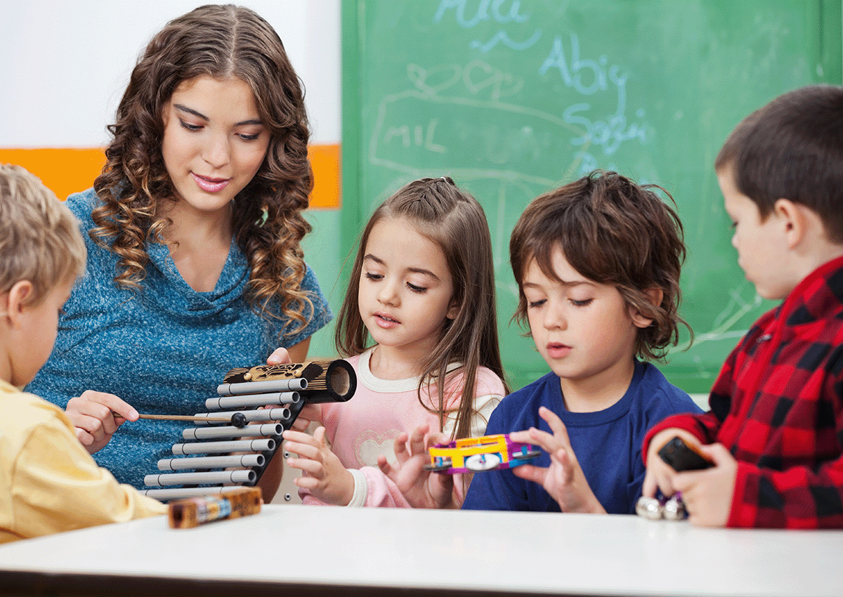 A teacher demonstrates using a musical instrument as students explore other instruments.
