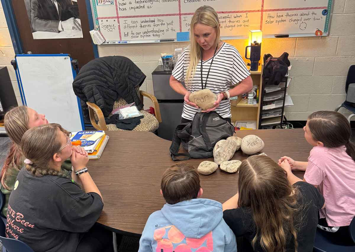 A teacher stands at a table, surrounded by students, holding a rock with fear written on it.
