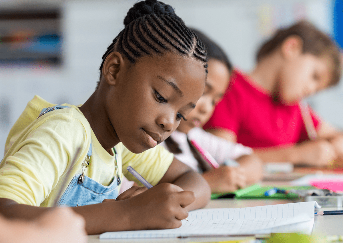 A young girl and her classmates are seated at a table, engaged in writing.