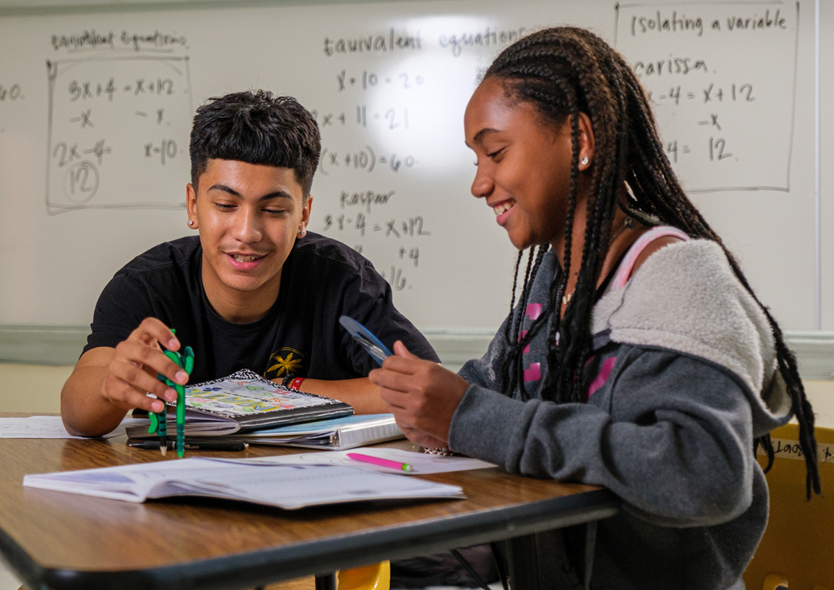 Two students are sitting with math tools in front of a whiteboard filled with equations.