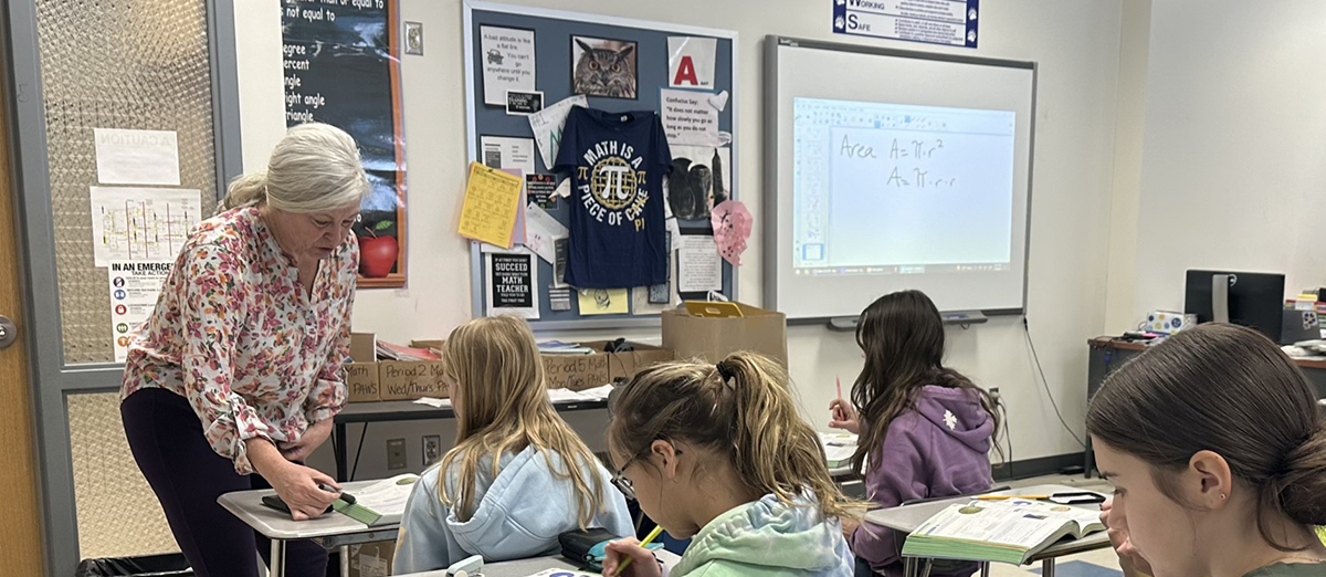 Students sitting at their desks write while a teacher checks in with a student.