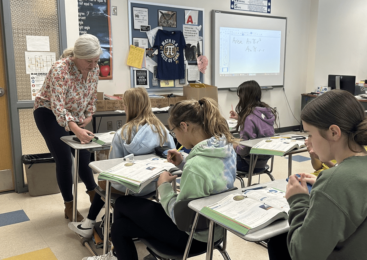 Students sitting at their desks write while a teacher checks in with a student.