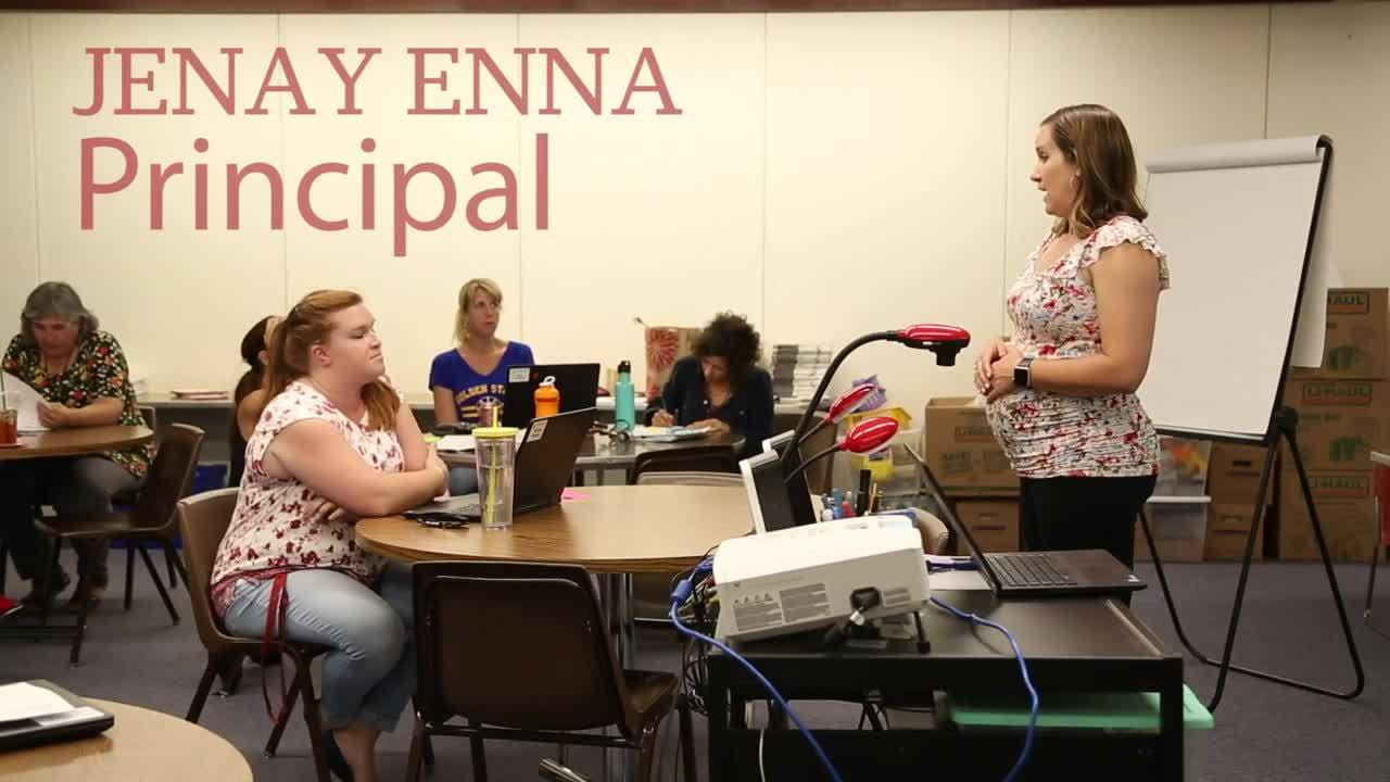 A principal stands at the front of a classroom, addressing seated staff members with laptops, papers, and a whiteboard visible in the background.