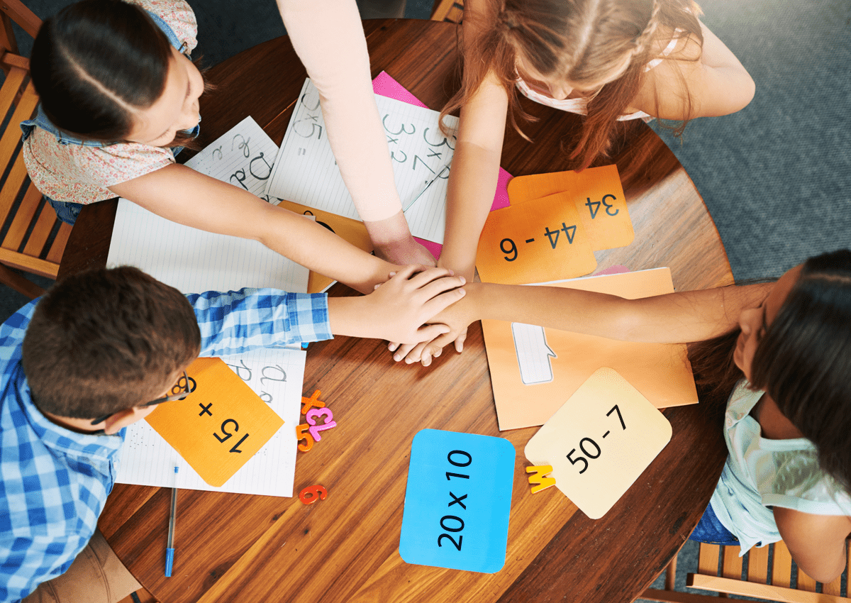Students stack hands while gathered around a table filled with math cards and notebooks.